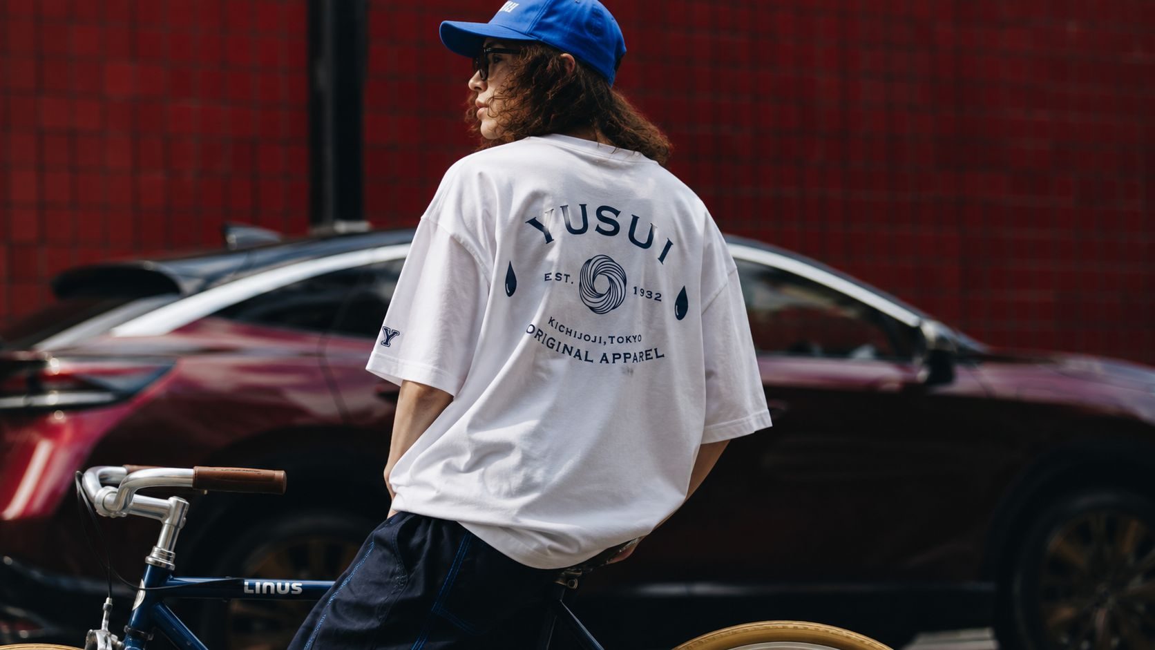 赤いタイル壁の前で、背中にYUSUIのアーチロゴが入った白Tシャツを着たモデル